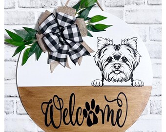 May include: A round wooden door hanger with a white top half and a brown bottom half. The top half features a black and white checkered bow with burlap accents and a black and white illustration of a Yorkshire Terrier peeking over the top. The bottom half has the word "Welcome" written in black cursive with a paw print in the center.