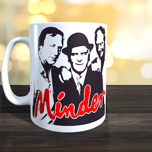 May include: White ceramic mug featuring a black and white image of three men. The men are wearing suits and hats. The word "Minder" is written in red letters across the bottom of the mug.