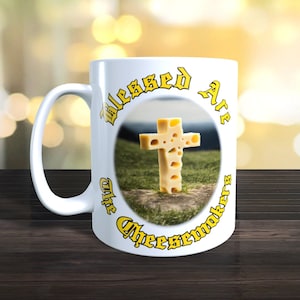 Könnte beinhalten: Weiße Keramiktasse mit einer gelben Grafik eines Kreuzes aus Schweizer Käse. Der Text um das Kreuz lautet "Blessed Are The Cheesemakers".