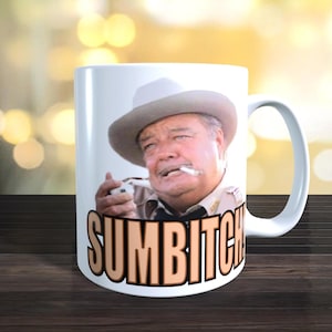 May include: White ceramic mug featuring a portrait of an older man in a cowboy hat, smoking a cigarette. The mug has the word "SUMBITCH" printed in large, orange letters across the front. The background is blurred.