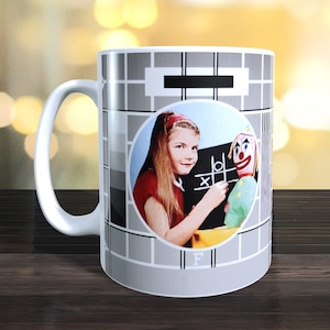 May include: White ceramic mug featuring a black and white television test pattern design. The pattern includes a circle with a girl playing a game with a clown doll. The letter "F" is visible at the bottom of the mug.