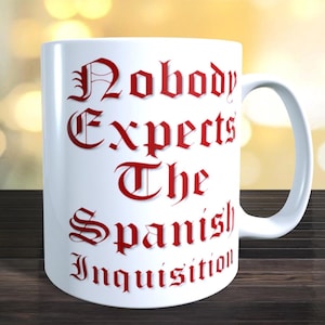 May include: White ceramic mug with the phrase "Nobody Expects The Spanish Inquisition" in red gothic lettering. The mug has a curved handle and is set against a blurred background.