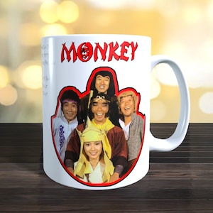 Könnte beinhalten: Weiße Keramik-Kaffeetasse mit rotem Rand und dem Wort "MONKEY" in roten Buchstaben. Die Tasse zeigt ein Foto der Besetzung der Fernsehserie "Monkey" aus den 1970er Jahren in Farbe.