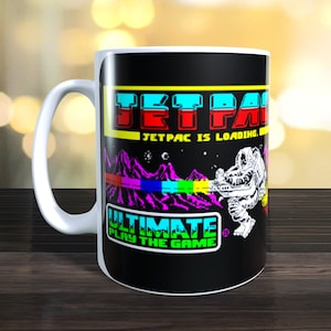 May include: A white ceramic mug with a black background featuring a retro video game design. The design includes the text "Jetpac is loading" and "Ultimate Play the Game".