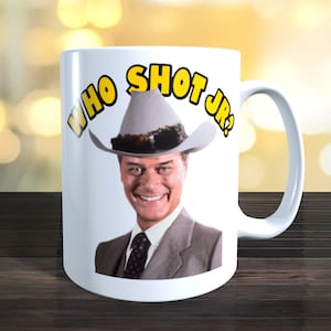 Op de afbeelding: Witte keramische koffiemok met een afbeelding van een man met een cowboyhoed en een pak. De tekst "WHO SHOT JR?" is in gele letters op de mok gedrukt.