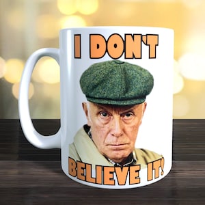 May include: White ceramic mug featuring a black and white image of a man wearing a green cap. The text "I DON'T BELIEVE IT!" is printed in orange on the mug.