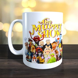 May include: White ceramic mug featuring a colourful image of the Muppets from "The Muppet Show" on the front. The text "The Muppet Show" is printed in yellow letters.