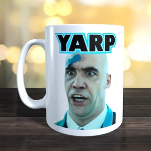 Może przedstawiać: Biały ceramiczny kubek do kawy z niebiesko-czarną grafiką twarzy mężczyzny z napisem "YARP" w niebieskich literach nad twarzą.