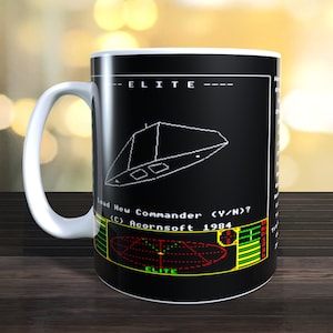 Puede incluir: Una taza de cerámica blanca con un diseño negro que presenta una nave espacial del clásico videojuego Elite. El diseño incluye el texto "ELITE" y "Load New Commander (Y/N)? (C) Acornsoft 1984".