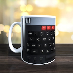 Pode incluir: Caneca de cerâmica branca com uma imagem preta e vermelha de um teclado de computador vintage. O teclado tem teclas etiquetadas "Escape", "Tab", "Caps Lock", "Ctrl", "Shift", "Z", "X", "C", "V", "F", "D", "S", "A", "Q", "W", "E", "R", "T", "Y", "U", "I", "O", "P", "[", "#", "1", "2", "3", "4", "5", "6", "7", "8", "9", "0", "-", "=", "Backspace", "Insert", "Home", "Page Up", "Delete", "End", "Page Down", "Arrow Up", "Arrow Left", "Arrow Down", "Arrow Right", "F1", "F2", "F3", "F4", "F5", "F6", "F7", "F8", "F9", "F10", "F11", "F12", "Print Screen", "Scroll Lock", "Pause Break", "Num Lock", "/