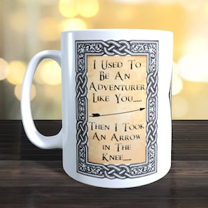 May include: White ceramic mug with a black and white design. The design features a Celtic knot border and text that reads "I used to be an adventurer like you..... Then I took an arrow in the knee....."