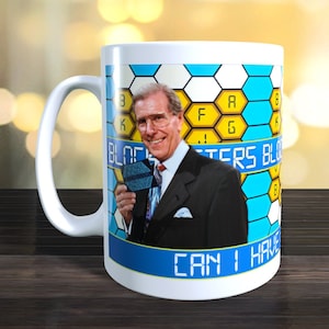 May include: White ceramic mug featuring a colourful image of a game show host holding a letter tile. The image includes the text "Blockbusters" and "Can I Have".