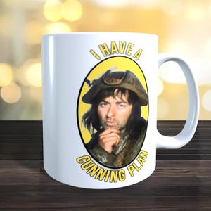 May include: White ceramic mug featuring a portrait of a man in a tricorn hat within a yellow oval. The text "I HAVE A" is above the portrait, and "A CUNNING PLAN" is below, all in yellow. The mug has a curved handle.