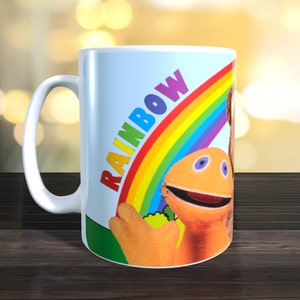 Könnte beinhalten: Weiße Keramiktasse mit einem Regenbogen-Design und dem Wort "RAINBOW" in Regenbogenfarben. Die Tasse zeigt auch eine Zeichentrickfigur mit orangefarbenem Gesicht und eine braune Figur. Die Tasse steht auf einer Holzoberfläche.