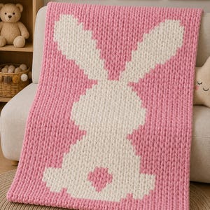 May include: A chunky knit blanket featuring a large white rabbit silhouette centred on a solid pink background. The blanket is constructed from thick, soft yarn with a visible, uniform loop texture throughout the entire rectangular surface.