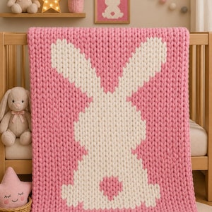 May include: A chunky knit blanket featuring a large white rabbit silhouette centred on a solid pink background. The blanket is constructed with thick yarn loops, creating a textured surface. The rabbit design includes a small pink heart detail.