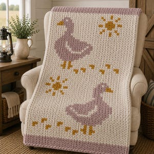 May include: A chunky knit throw blanket in cream, featuring two mauve ducks and two mustard yellow suns. The blanket has a thick mauve border at the top and bottom, with small mustard yellow footprints trailing behind each duck across the surface.