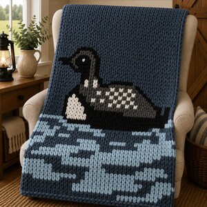 May include: A chunky knit throw blanket in deep navy blue featuring a large, centered graphic of a loon swimming on water. The loon is rendered in black, grey, and white yarn, while the water below is depicted in varying shades of light blue.