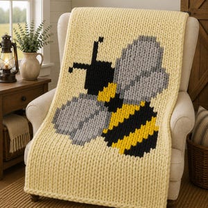 Könnte beinhalten: Eine grob gestrickte Wohndecke in Hellgelb mit einer großen, mittigen Bienen-Grafik. Das Bienendesign besteht aus schwarzem, gelbem und grauem Garn und zeigt einen gestreiften Körper, einen schwarzen Kopf mit Fühlern sowie graue Flügel.