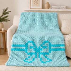 May include: A chunky knit throw blanket in a light turquoise color featuring a prominent bow design in a darker shade of turquoise. The blanket is crafted with thick, soft yarn loops and is draped over a neutral-colored sofa, showcasing its texture.