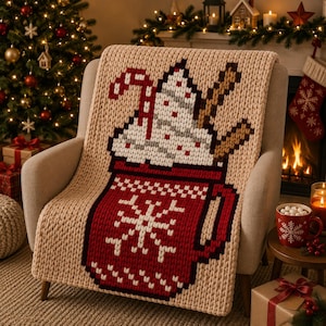 May include: A chunky knit throw blanket in beige featuring a large pixelated graphic of a red mug filled with whipped cream, a candy cane, and a cinnamon stick. The red mug displays a white snowflake pattern and a checkered border design.