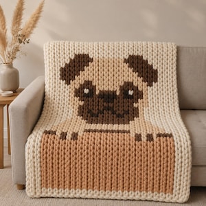 May include: A chunky knit throw blanket featuring a large, centered pug dog face design. The blanket is primarily cream-colored with dark brown accents for the pug's ears, eyes, and muzzle, and a light tan section at the bottom representing paws.