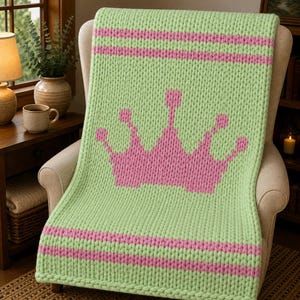 Puede incluir: Una manta de punto grueso en color verde claro con un gráfico de corona rosa grande en el centro. La manta tiene dos rayas horizontales rosas cerca de los bordes superior e inferior, creadas con hilo grueso en un patrón de punto texturizado.