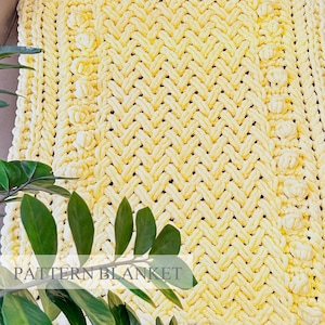 May include: A pale yellow chunky knit blanket featuring a complex braided cable pattern and vertical rows of raised bobble stitches. The thick yarn creates a textured surface with a repeating geometric design throughout the entire fabric.