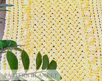 May include: A pale yellow chunky knit blanket featuring a complex braided cable pattern and vertical rows of raised bobble stitches. The thick yarn creates a textured surface with a repeating geometric design throughout the entire fabric.
