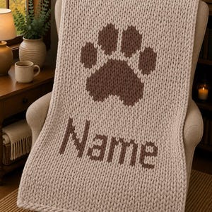 May include: A chunky knit throw blanket in a light beige color featuring a large dark brown paw print design at the top and the word Name in dark brown letters centered below the paw print, all created with a thick yarn texture.