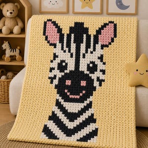 Pode incluir: Uma manta de malha grossa na cor amarelo-pálido com um gráfico grande de uma zebra centralizado. A zebra é composta por pontos de fio preto e branco com detalhes em rosa no interior das orelhas e um pequeno sorriso rosa bordado no rosto.