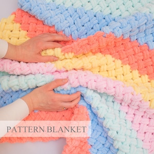Op de afbeelding: Een grof gebreide deken met een zacht, gestructureerd patroon met diagonale strepen in pasteltinten van lichtblauw, perzik, geel en roze. Het textiel heeft een dikke, geluste constructie en bevat de tekst PATTERN BLANKET onderaan.