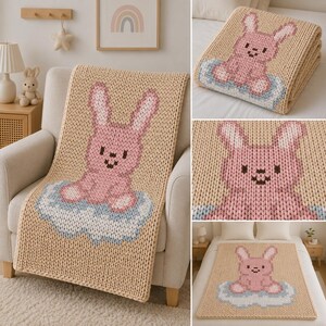 May include: A chunky knit throw blanket in a light beige color featuring a large, centered graphic of a pink rabbit sitting on a light blue cloud. The rabbit has dark brown eyes and a dark brown nose, with white accents on its ears and feet.