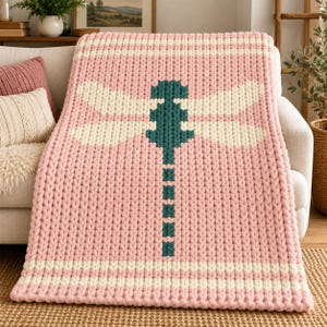 May include: A chunky knit throw blanket featuring a large, centered dragonfly motif. The blanket is primarily light pink with a dark teal dragonfly body and cream-colored wings. Horizontal cream stripes are visible at the top and bottom edges.