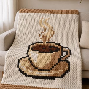 May include: A chunky knit throw blanket featuring a pixel art design of a coffee cup on a saucer. The blanket is primarily cream-colored with brown borders at the top and bottom. The coffee cup graphic uses shades of brown, tan, and white.