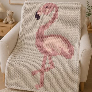 Könnte beinhalten: Eine grob gestrickte Wohndecke in Cremefarbe mit einem großen, verpixelten Flamingomotiv in Rosa-, Hellrosa- und Dunkellila-Tönen. Die Decke hat eine dicke, strukturierte Garnkonstruktion mit einem durchgehend sichtbaren Maschenmuster.