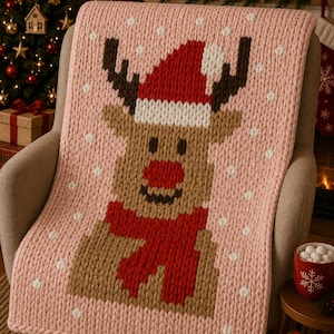 May include: A chunky knit blanket in light pink featuring a large reindeer graphic. The reindeer has brown fur, a bright red nose, a red scarf, and a red Santa hat with a white pom-pom. Small white dots are scattered across the pink background.