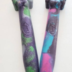 May include: Two Crayola brand crayons with a flower-shaped top. One crayon is purple, green, and black with glitter. The other crayon is pink, blue, and black.