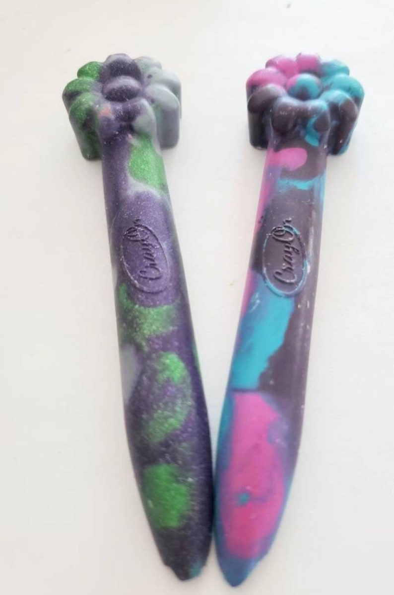 May include: Two Crayola brand crayons with a flower-shaped top. One crayon is purple, green, and black with glitter. The other crayon is pink, blue, and black.