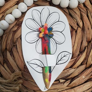 May include: A crayon flower craft on a white paper cutout. The flower's petals are made of colorful melted crayons. The stem is a crayon with the word "flower" visible. The background is a woven texture with white beads.