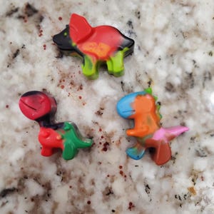 May include: Three colorful dinosaur-shaped crayons. The crayons are red, green, black, orange, blue, and pink.