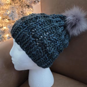 May include: A dark teal crocheted beanie with a gray faux fur pom-pom. The hat is displayed on a white foam head. The hat has a textured stitch pattern and is a winter accessory.