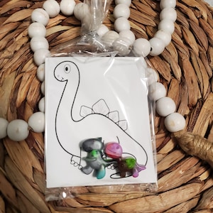 May include: A clear plastic bag containing a set of four small dinosaur figurines. The dinosaurs are in various colors, including green, pink, and blue. The bag also contains a black and white coloring page of a dinosaur.