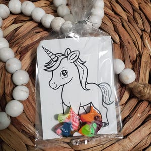 May include: A white card with a black and white line drawing of a unicorn. Three small unicorn-shaped crayons are in front of the card. The crayons are multi-colored and have a rainbow design.