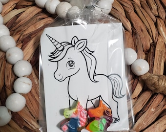 May include: A white card with a black and white line drawing of a unicorn. Three small unicorn-shaped crayons are in front of the card. The crayons are multi-colored and have a rainbow design.