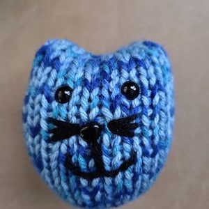 May include: A knitted blue cat face with black embroidered details. The cat has two black button eyes, a black nose, and a smiling mouth. The yarn is a mix of light and dark blue shades.