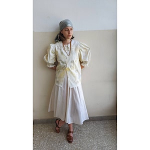 May include: A woman wearing a white blouse with a lace collar and a yellow and blue tie-dye pattern. She is also wearing a white skirt and a blue bandana on her head.