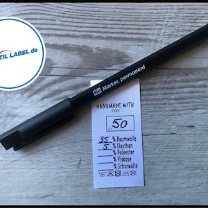 Könnte beinhalten: Ein weißes Etikett mit schwarzem Text, der "HANDMADE WITH DOVE" und "Gr. 50" lautet. Das Etikett listet auch die Stoffzusammensetzung auf: 95% Baumwolle, 5% Elasthan, und enthält Pflegehinweise. Ein schwarzer Permanentmarker liegt auf dem Etikett.