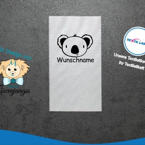 Könnte beinhalten: Ein weißes Stofflabel mit einer schwarz-weißen Koalabären-Illustration. Der Text "Wunschname" ist unter dem Koala gedruckt. Der Text "Textil Label" ist in einem blauen Kreis auf der rechten Seite des Labels gedruckt. Der Text "Unsere Textiletiketten Ihr Textiletikett" ist unter dem Kreis gedruckt.
