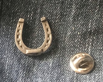 Könnte beinhalten: Silberner Hufeisen-Anhänger mit einer Sicherheitsnadel. Das Hufeisen ist mit einem strukturierten Design verziert.